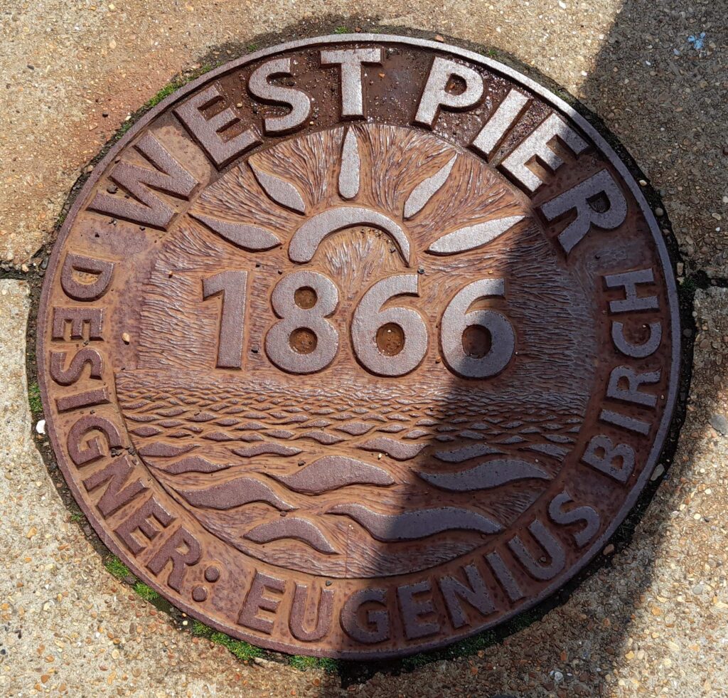 Round iron plaque embedded in concrete, with the words 'West Pier, 1866, Designer: Eugenius Birch'.
